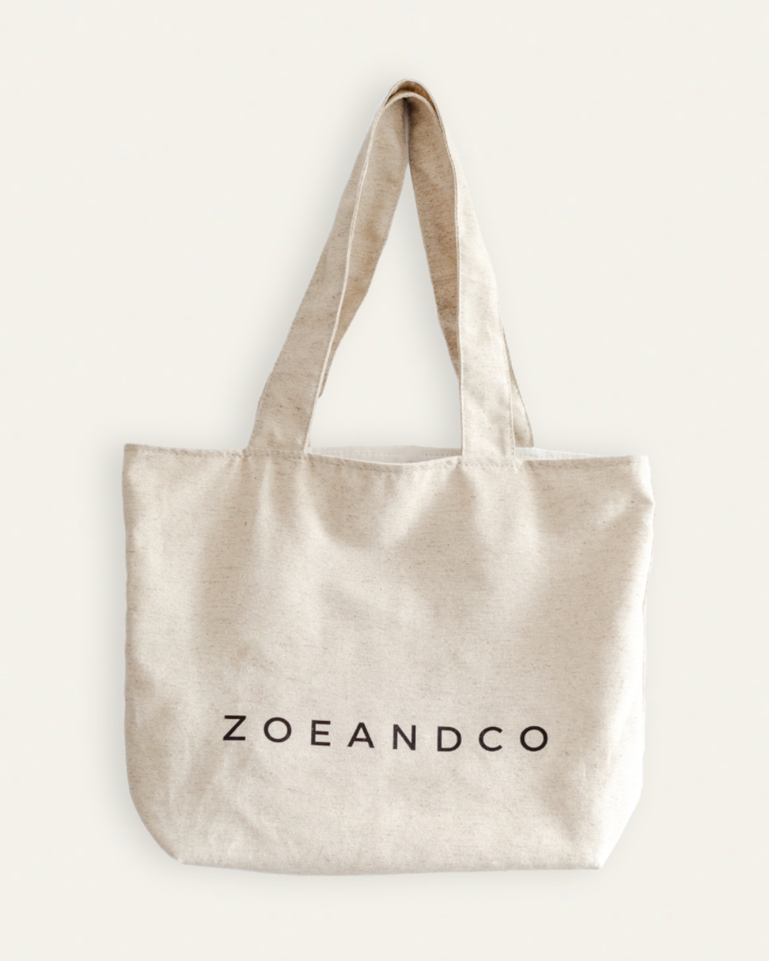 A natural beige canvas tote bag with black printed text reading 'ZOEANDCO' on the front, displayed against a solid off-white background.