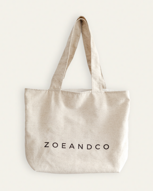 A natural beige canvas tote bag with black printed text reading 'ZOEANDCO' on the front, displayed against a solid off-white background.